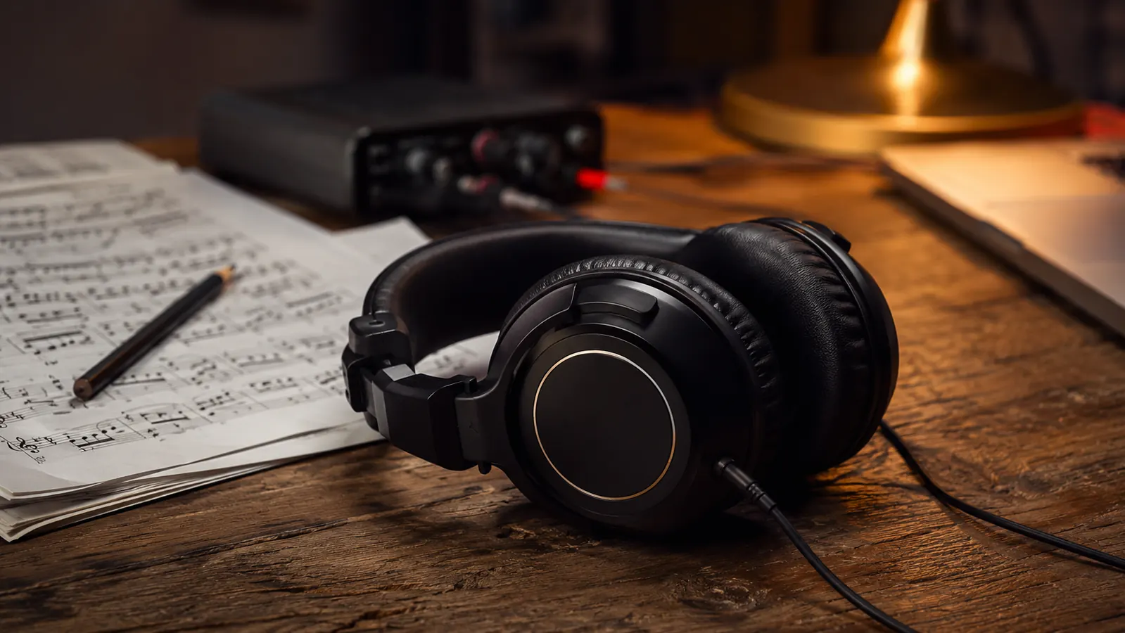 Professional studio monitor headphones resting on a worn wooden teaching desk beside sheet music and a small audio interface.
